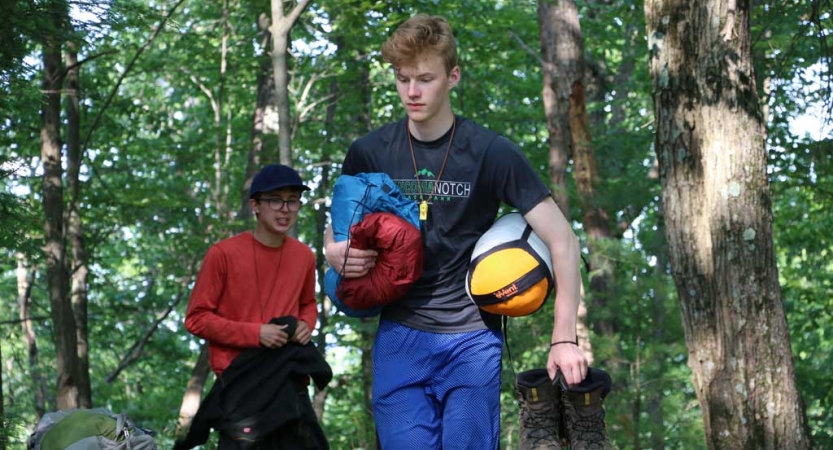Two people carry gear through a wooded area